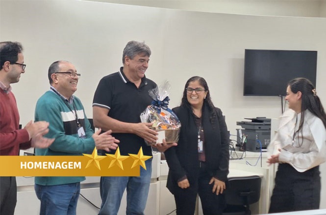Na imagem, há cinco pessoas em uma sala, parecendo participar de uma homenagem. Uma pessoa no centro está segurando uma cesta de presente, enquanto as outras parecem estar aplaudindo ou sorrindo. No canto inferior esquerdo, há um gráfico com a palavra "HOMENAGEM" seguida por três estrelas. Ao fundo, é possível ver alguns equipamentos de escritório, como uma televisão e mesas com cadeiras. A atmosfera parece ser de celebração ou reconhecimento.