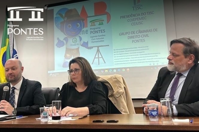 A imagem mostra três pessoas sentadas à mesa, participando de algum tipo de evento ou reunião. Eles estão vestidos formalmente, sugestivos de um ambiente profissional. Atrás deles, há uma tela projetada com informações do Programa PONTES, sugerindo uma iniciativa relacionada a soluções humanizadas para famílias. As bandeiras do Brasil e outra instituição estão visíveis ao lado esquerdo da tela.