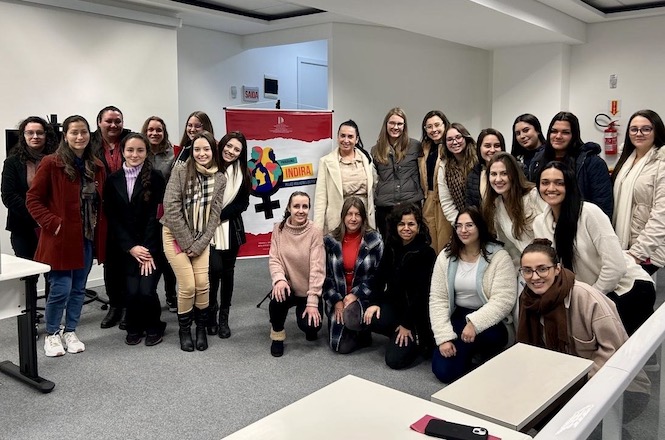 A imagem mostra um grupo de mulheres em uma sala. Elas estão posando para uma foto em frente a uma faixa colorida que parece ter o símbolo de igualdade e as palavras "Indira" e "Agora". As mulheres estão sorrindo e usam roupas de inverno. Há mesas e cadeiras na sala, sugerindo que pode ser um ambiente educativo ou de reunião.