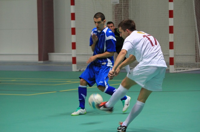A imagem mostra um jogo de futsal com dois jogadores em quadra. Um jogador vestido de azul está à esquerda, observando o adversário. O outro jogador, vestido de branco, está à direita, fazendo um movimento de chute na bola. Ao fundo, é possível ver o gol e a rede da quadra.