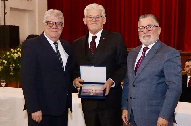 Na imagem, há três homens em um ambiente formal. Eles estão vestidos com ternos e gravatas. O homem do meio segura uma placa ou prêmio. Ao fundo, há uma cortina vermelha e uma mesa com toalha branca. Parece ser um evento de premiação ou reconhecimento.