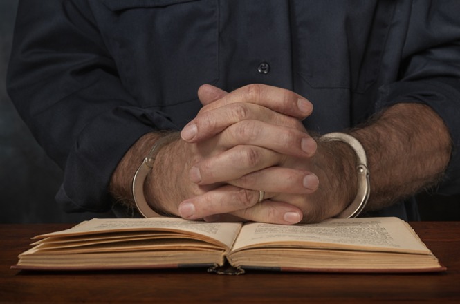 A imagem mostra as mãos de uma pessoa algemada, cruzadas sobre um livro aberto. A pessoa está usando uma roupa escura, e a cena parece representar uma situação de reflexão ou oração enquanto está algemada.