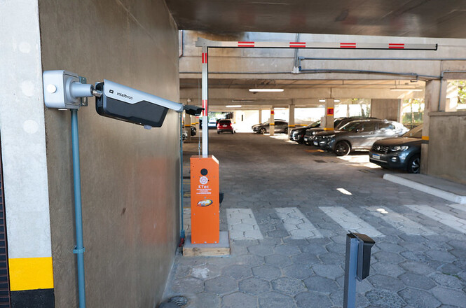 A imagem mostra a entrada de um estacionamento coberto. À esquerda, há uma câmera de vigilância instalada na parede, e ao lado dela há uma cancela automática laranja com um dispositivo de controle de acesso. O piso é de blocos de concreto. Mais ao fundo, há vários carros estacionados, e há colunas de concreto sustentando o teto. A iluminação é natural e decorre da luz do dia.