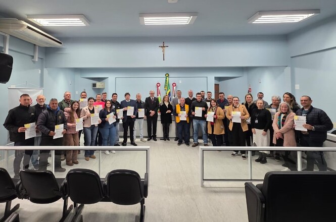 A imagem mostra um grupo de pessoas posando para uma foto em um ambiente que parece ser uma sala de reuniões ou auditório. Todos estão em pé, muitos segurando papéis nas mãos. Atrás deles, há um emblema ou brasão na parede. O espaço tem cadeiras à frente, de costas para a câmera, e é decorado de forma simples. Parece ser um evento formal ou cerimônia.