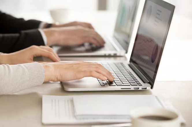 A imagem mostra duas pessoas trabalhando em laptops. Há gráficos exibidos nas telas dos computadores. Também é possível ver papéis anotados sobre a mesa e uma xícara de café. As mãos das pessoas estão sobre os teclados, sugerindo que estão digitando ou interagindo com o computador. O ambiente parece ser um espaço de trabalho colaborativo ou um escritório.