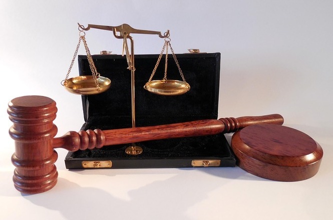 A imagem mostra um martelo de juiz e uma balança de justiça. O martelo é de madeira e está posicionado em frente a uma balança dourada, que está sobre um fundo branco. Esses itens são frequentemente associados ao sistema judicial e simbolizam a justiça e a autoridade legal. A balança está dentro de uma caixa preta aberta.