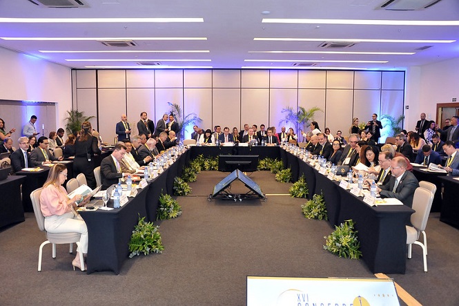 A imagem retrata uma conferência ou reunião em um ambiente interno, provavelmente em uma sala de conferências. No primeiro plano, há várias mesas dispostas em forma de 'U', cobertas por toalhas de mesa pretas e decoradas com plantas verdes. As mesas estão cheias de pessoas bem vestidas em trajes formais, sugerindo um encontro profissional ou de negócios.  Os participantes estão sentados ao redor das mesas, muitos deles envolvidos em leitura ou anotação de documentos. Alguns olham para a frente, possivelmente ouvindo um orador. Há também várias garrafas de água e copos sobre a mesa, indicando que a reunião pode ser longa.  O fundo da imagem mostra um painel grande, com iluminação que transmite uma atmosfera formal e organizada. Plantas estão posicionadas ao longo das paredes, contribuindo para a ambientação. Observam-se pessoas em pé nas laterais, que podem ser equipe de apoio ou segurança.  A emoção transmitida é de seriedade e concentração, típica de eventos corporativos importantes. O clima sugere uma atmosfera de discussão intensa ou apresentação de tópicos relevantes.  A iluminação interna é clara, típica de um local fechado sem janelas visíveis, o que dificulta identificar a hora do dia ou a estação do ano. A aparência é totalmente realista, sem evidências de ser uma imagem desenhada, animada ou gerada por computador.