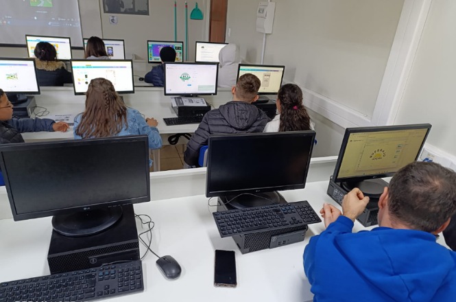 A imagem retrata uma sala de aula de informática.   No primeiro plano, há computadores sobre uma mesa branca, com teclados e mouses. No fundo, outras pessoas estão usando computadores adicionais.  Os principais objetos são os computadores e seus periféricos, como teclados e mouses. As pessoas presentes estão sentadas em frente aos computadores, aparentemente focadas em suas atividades.  As pessoas parecem estar concentradas em suas telas, sugerindo que estão trabalhando ou aprendendo algo no computador.  A atmosfera é provavelmente de estudo ou trabalho, transmitindo concentração e foco.  A sala parece ser uma sala de aula ou um laboratório de informática, equipada para o ensino ou trabalho colaborativo em um ambiente educacional.  Não há indicações visíveis do horário ou da estação na imagem, uma vez que é em um ambiente interno.  A imagem é realista e fotografada, capturando uma cena cotidiana em um ambiente escolar ou de treinamento.