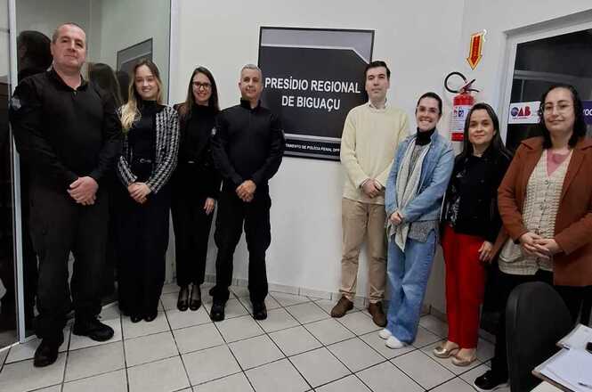 Observa-se um grupo de pessoas posando juntas em um ambiente interno. Ao fundo, há uma parede clara com um quadro que diz "Presídio Regional de Biguaçu". O piso é composto por azulejos claros. A cena é composta por várias pessoas vestidas de maneira formal ou semi-formal; algumas estão trajando roupas escuras, enquanto outras usam tons claros. Há também uma placa na parede e um extintor de incêndio. As pessoas estão de pé, lado a lado, possivelmente para uma foto de grupo. Elas parecem estar em uma postura amigável e formal, sugerindo um ambiente profissional. A expressão geral dos participantes é neutra, sugerindo um ambiente de trabalho ou uma ocasião oficial. A atmosfera parece ser a de uma reunião ou evento institucional. O ambiente parece ser o interior de uma instituição pública, possivelmente uma área administrativa ou de recepção de um presídio, dado o texto na parede.Não há indicações claras do tempo do dia ou da estação do ano, pois a cena está em um ambiente fechado e sem janelas visíveis. A imagem é uma fotografia realista, sem elementos de desenho, animação ou geração por computador.