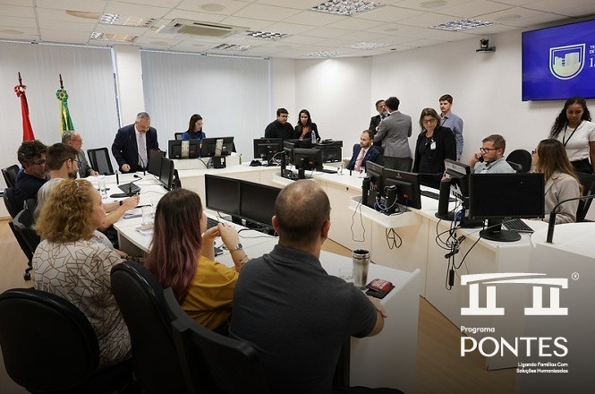 A imagem retrata uma sala de conferências onde várias pessoas estão participando de uma reunião. No primeiro plano, há algumas pessoas sentadas em cadeiras ao redor de mesas equipadas com computadores e monitores. Elas parecem atentas, possivelmente discutindo ou ouvindo uma apresentação. No fundo, mais pessoas estão de pé, parecendo estar envolvidas em interações menores ou observando o que está acontecendo na sala. Algumas parecem estar reunidas em pequenos grupos. Os objetos principais incluem mesas, cadeiras, computadores, monitores e algumas bandeiras, possivelmente indicando que a sala pertence a um ambiente institucional ou governamental. As pessoas parecem estar envolvidas em atividades relacionadas a trabalho ou discussões importantes. A atmosfera da imagem sugere um ambiente profissional e colaborativo. As expressões das pessoas indicam concentração e engajamento, típicas em reuniões de trabalho ou conferências. O local aparente é uma sala de conferências moderna, possivelmente em um edifício governamental ou de uma empresa. O interior é bem iluminado, indicando que a imagem pode ter sido capturada durante o dia. O estilo é realista, e a foto parece ter sido tirada em um ambiente moderno e funcional. A presença de um logotipo no canto inferior direito da imagem, juntamente com a tela ao fundo exibindo um escudo, sugere que se trata de um evento ou programa específico chamado "Programa Pontes".