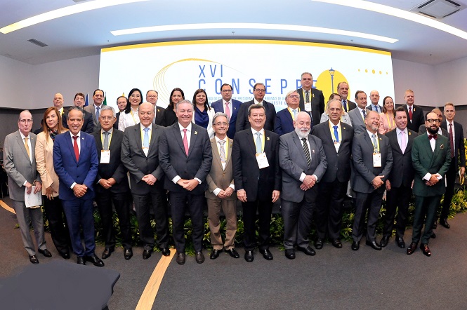 A imagem apresenta um grupo de pessoas reunidas em um auditório. No primeiro plano, há várias pessoas em pé, provavelmente posando para uma foto oficial. Todas estão vestidas de forma formal, usando ternos e gravatas, sugerindo um evento profissional ou conferência. Algumas pessoas têm crachás, indicando que são participantes de um evento.No fundo, pode-se ver um grande painel com a inscrição "XVI CONSEP", que parece ser o nome do evento, possivelmente uma conferência ou seminário. O painel tem um design simples com cores brancas, amarelas e azuis.As pessoas no grupo aparentam estar interagindo de maneira cordial, possivelmente sorrindo ou mantendo uma expressão neutra, o que sugere um ambiente formal, mas amigável. A atmosfera é profissional e organizada, típica de eventos corporativos.O local parece ser um espaço de conferência moderno e bem iluminado, com iluminação artificial clara. Embora a hora exata do dia não seja evidente, a iluminação sugere que a foto foi tirada em um ambiente interno durante o dia.A imagem é realista e parece ser uma fotografia, capturando um momento específico em um contexto profissional.