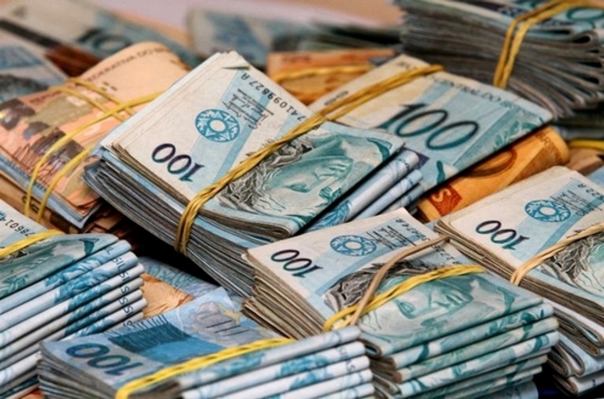 A imagem retrata várias pilhas de notas de dinheiro no que parece ser uma superfície de mesa. No primeiro plano, há maços de notas organizados e amarrados com elásticos. As notas são de valores variados, mas as de cem reais, em destaque, são mais visíveis. A presença de várias notas amontoadas uma sobre a outra transmite a ideia de uma quantia significativa de dinheiro.No plano de fundo, outras pilhas de notas são visíveis, mas com menos nitidez, sugerindo que a área está cheia de dinheiro, possivelmente em um escritório ou um ambiente financeiro. Os maços de notas dão a impressão de organização e grandeza, talvez associada a uma transação ou coleta de dinheiro.
