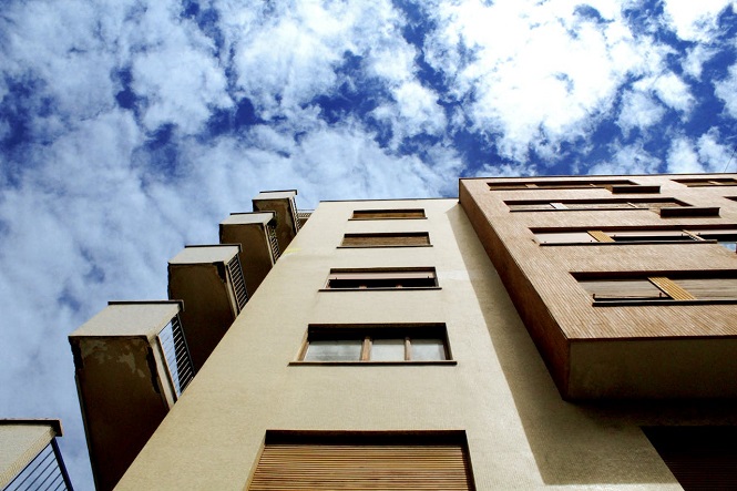 A imagem mostra a fachada de um edifício visto de uma perspectiva baixa, olhando para cima. No primeiro plano, temos a estrutura do prédio, que ocupa a maior parte da imagem. Ao fundo, podemos observar um céu azul com várias nuvens brancas dispersas, criando um contraste interessante com o edifício.