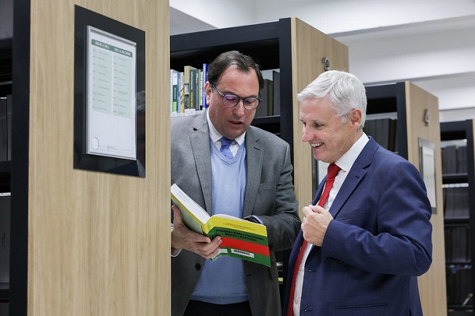 Foto horizontal: Francisco Oliveira Neto lê livro com José Igreja Matos, presidente do TRP, em biblioteca, tons claros.