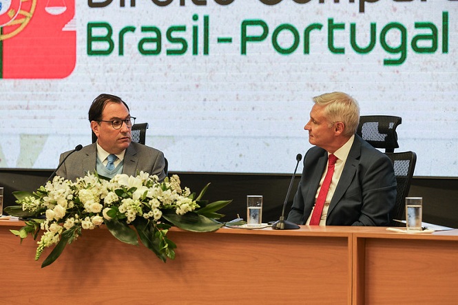 A imagem apresenta um cenário em uma conferência ou reunião formal. No primeiro plano, há duas pessoas sentadas em uma mesa grande de madeira. Eles estão vestindo ternos, ressaltando a formalidade do evento. À esquerda, um homem está olhando para o outro, possivelmente envolvidos em uma discussão ou aguardando sua vez para falar. À direita, o outro homem parece estar olhando para o primeiro, indicando que estão interagindo entre si.Na mesa há um arranjo de flores brancas, adicionando um toque de elegância ao ambiente. Há também microfones e copos de água em frente a cada homem, típicos de conferências. O fundo da imagem mostra um painel com as palavras "Brasil-Portugal", sugerindo que o evento está relacionado a colaboração ou palestras entre esses dois países. O painel é parte de uma tela grande com gráficos, reforçando o caráter formal e possivelmente internacional do evento.