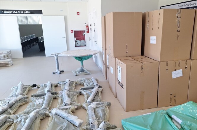 A imagem mostra uma sala com vários itens embalados. No primeiro plano, há caixas grandes de papelão empilhadas à direita. Em frente a elas, alguns objetos longos e volumosos estão embrulhados em plástico transparente, dispostos no chão. Também há uma mesa coberta com plástico verde.No plano de fundo, vê-se uma porta aberta que leva a uma sala adjacente com cadeiras alinhadas, sugerindo que é uma sala de tribunal ou conferências, indicada pela placa "TRIBUNAL DO JÚRI" acima da porta. Outro detalhe visível é um extintor de incêndio e rolos de papel ou tecido próximos à parede.