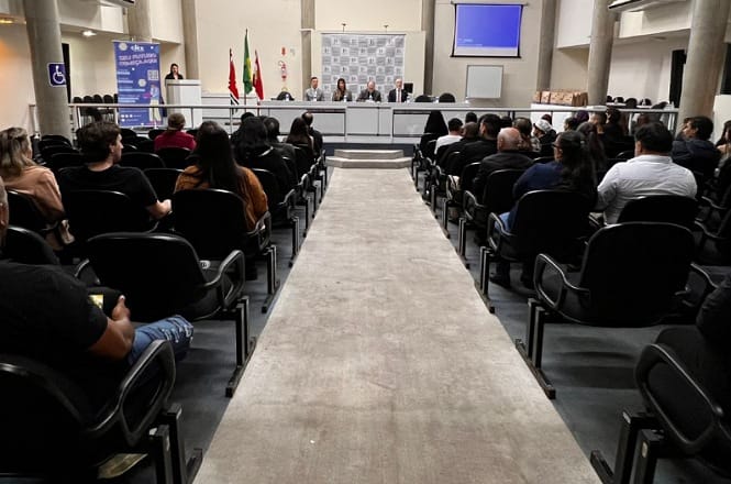 A imagem mostra o interior de uma sala de conferências ou auditório. No primeiro plano, pode-se ver fileiras de cadeiras ocupadas por pessoas sentadas, aparentemente assistindo a um evento ou apresentação.No fundo da imagem, há uma mesa comprida com diversas pessoas sentadas, que parecem ser palestrantes ou membros de um painel. Atrás deles, há algumas bandeiras e um banner indicativo, além de uma tela projetada ou um quadro branco, possivelmente para apresentações.