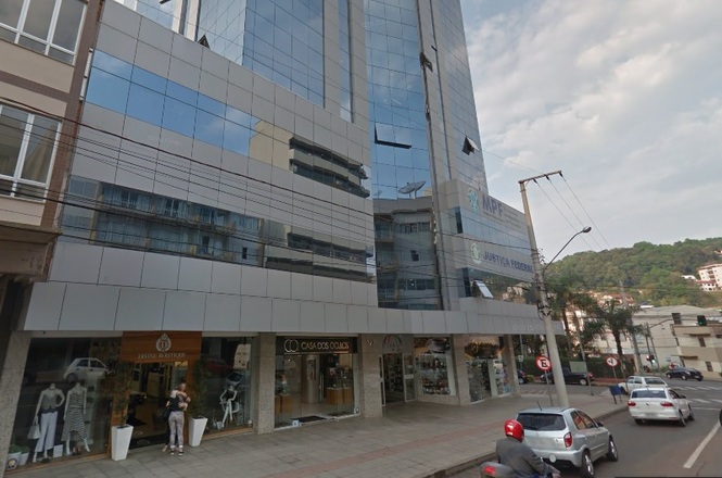 A composição da cena é de uma rua urbana, onde o foco principal é um edifício alto de múltiplos andares. O edifício tem um design moderno e está cercado por lojas e edifícios menores. O foco principal é o edifício alto com fachada de vidro e paredes cinza claro, bem como várias lojas ao longo da rua. A estrutura de composição enfatiza a altura do edifício, com o fundo ocupado por ele e elementos menores à sua frente. As lojas parecem estar voltadas para a rua, e suas janelas de loja exibem a visão de dentro. A movimentação é pequena, com apenas alguns pedestres visíveis e um motociclista no plano de fundo.