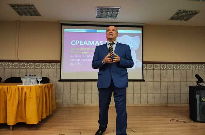 Um homem de terno azul marinho está em uma apresentação, provavelmente em uma sala de conferências. Há um projetor e uma tela exibindo um título relacionado a assuntos de apoio a crianças. Uma mesa com toalhas amarelas e copos com água fica à frente, na frente da platéia.