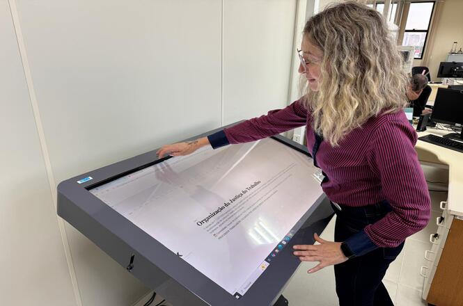 Na imagem, vemos uma mulher interagindo com uma grande tela sensível ao toque, colocada na diagonal em uma espécie de suporte. Ela parece estar usando a tela de forma profissional, possivelmente em um ambiente de escritório. A mulher está usando óculos e parece estar concentrada na tela. Está vestida com um suéter listrado em tons de bordô. Ela está interagindo diretamente com a tela, possivelmente navegando ou manipulando o conteúdo exibido.
