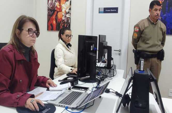 Uma sala de escritório com pessoas em frente a computadores.  Há mulheres usando  jaquetas e um homem com uniforme de policial militar. Há vários computadores e laptops na mesa.  Há um quadro na parede com um design abstrato.