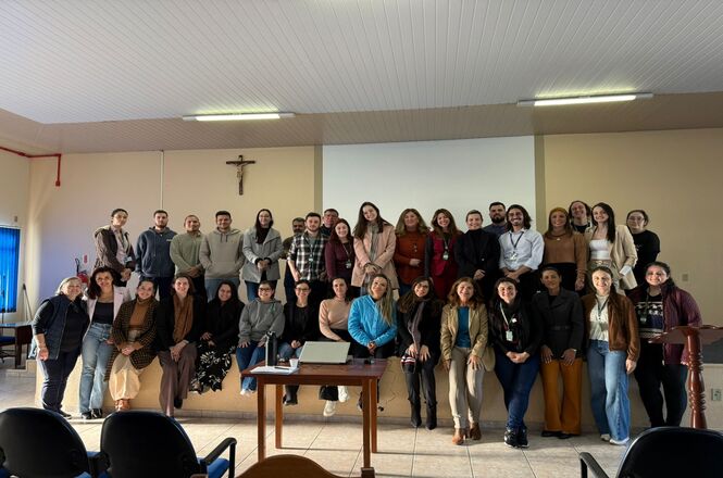 Um grupo grande de pessoas está reunido em uma sala, possivelmente em uma instituição educacional ou profissional.  A maioria das pessoas está em pé, posicionadas em várias fileiras, e algumas estão sentadas em uma pequena elevação. A sala é clara e bem iluminada, com paredes de cor clara e teto revestido. Há uma cruz na parede atrás do grupo. A imagem sugere um evento de grupo, como uma reunião, workshop ou formação.