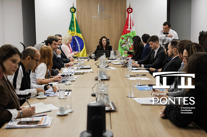 Vários indivíduos estão sentados em torno de uma mesa comprida, possivelmente em uma reunião. As bandeiras do Brasil e de outro país estão atrás da mesa. Há diversos documentos e itens na mesa, além de copos e garrafas d'água. Os participantes estão vestidos com roupas formais, indicando um ambiente de trabalho ou reunião oficial.  Há também computadores portáteis na mesa.