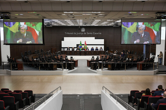 Interior de um tribunal, possivelmente o Tribunal de Justiça de Santa Catarina, mostrando a área de audiências.  Várias pessoas estão sentadas em cadeiras, tanto na área central como nas arquibancadas. Há uma mesa grande no centro, com pessoas sentadas, e duas telas grandes exibem imagens.  A decoração do ambiente é formal e austera.