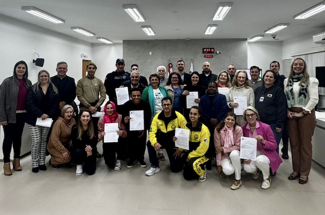 Grupo de pessoas, possivelmente participantes de um curso ou treinamento, em um ambiente institucional.  Várias pessoas estão em pé e outras estão ajoelhadas, segurando documentos. O ambiente é claro e parece ser uma sala de conferências ou auditório.