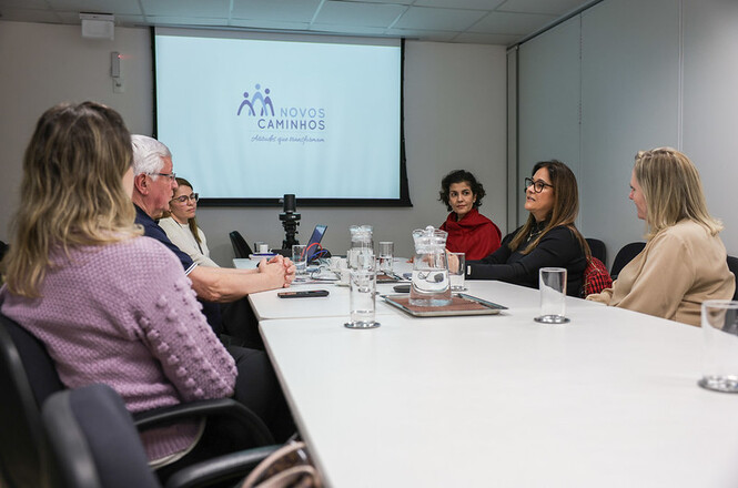 A imagem mostra uma reunião em uma sala de conferências. No primeiro plano, vemos uma mesa branca ao redor da qual estão sentadas várias pessoas, um grupo misto de homens e mulheres, que parecem estar engajados em uma discussão ou reunião. A maioria deles está olhando na direção de um quadro ou tela. No fundo, há uma tela de projeção com o logo "Novos Caminhos" e a frase "Educar para transformar", sugerindo que a reunião pode estar relacionada a iniciativas educacionais ou sociais. A presença de um projetor sobre a mesa sugere a apresentação de informações ou compartilhamento de conteúdo visual durante a reunião.