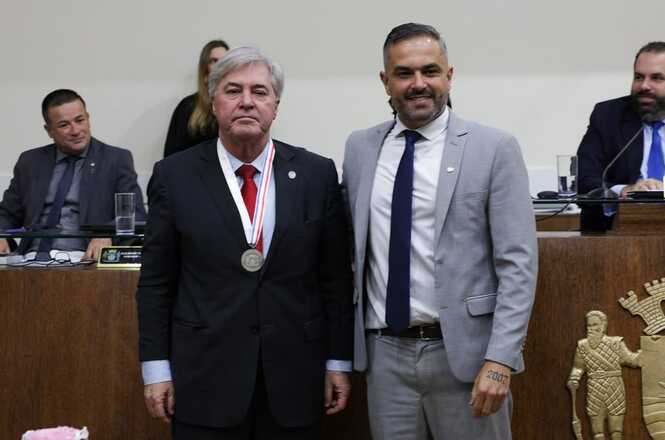 A imagem mostra duas pessoas em destaque no primeiro plano, posando juntas para uma foto. O homem à esquerda está vestindo um terno escuro com gravata vermelha e uma medalha pendurada no pescoço. O homem à direita também está de terno, mas de cor cinza, com camisa branca e gravata azul escura. Ambos parecem estar em um ambiente formal, possivelmente uma cerimônia de premiação ou evento oficial.