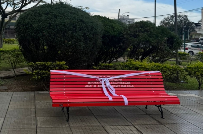 No primeiro plano, há um banco de parque pintado de vermelho. O banco está decorado com uma fita branca, que está amarrada formando um laço. No encosto do banco, há uma mensagem escrita, que parece apresentar um texto de conscientização sobre a violência contra as mulheres.