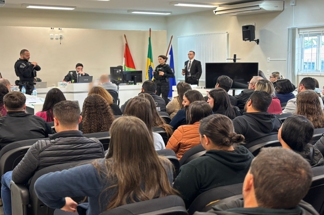 A imagem mostra uma sala de audiência ou reunião. No primeiro plano, há várias pessoas sentadas, de costas, aparentemente assistindo ou participando de um evento. A presença de juízes, advogados ou policiais sugere que pode ser um tribunal ou lugar de trabalho relacionado ao sistema judicial.