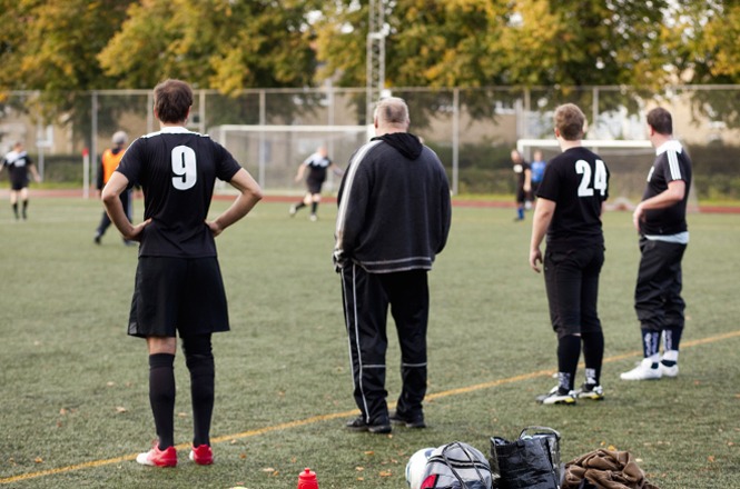 A imagem mostra um cenário de jogo de futebol. No primeiro plano, há quatro pessoas em pé à beira do campo. Três delas vestem uniformes de futebol com os números 9, 24 e outro uniforme sem número visível. Um dos indivíduos, diferente dos outros, parece não vestir uniforme, sugerindo que pode ser um técnico ou observador. No fundo, é possível ver outros jogadores em ação no campo, indicando que o jogo está em andamento. As pessoas no primeiro plano parecem estar assistindo ao jogo ou esperando para entrar em campo. O ambiente transmite uma atmosfera de concentração e expectativa, típica de um evento esportivo. Os jogadores parecem estar focados no jogo. O cenário é um campo de futebol ao ar livre, com grama sintética, cercado por árvores que têm folhas em tons que sugerem ser outono. A iluminação natural e a presença de árvores indicam que a cena ocorre durante o dia. A imagem é realista e parece ser uma fotografia contemporânea.