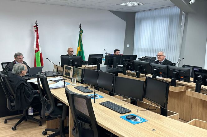 A imagem mostra uma sala de tribunal ou conferência. No fundo, há bandeiras, possivelmente simbolizando nacionalidade ou estado, posicionadas próximas a uma parede branca. A cena é composta por várias pessoas sentadas em torno de uma mesa de formato retangular com monitores de computador. Cada pessoa está em frente a um computador. Os objetos principais incluem computadores, papéis e microfones. As pessoas estão sentadas, provavelmente participando de uma reunião ou julgamento. A configuração sugere uma atmosfera formal e profissional. A imagem transmite uma sensação de seriedade e foco, típico de um ambiente de trabalho especializado. A sala parece ser de uma instituição governamental ou judiciária, indicada pelas bandeiras e pela configuração formal. Não há luz natural visível que indique a hora do dia. A iluminação artificial sugere que pode ser em qualquer momento do dia. Não há indícios do clima ou estação. É uma fotografia realista, sem elementos de desenho, animação ou geração por computador.