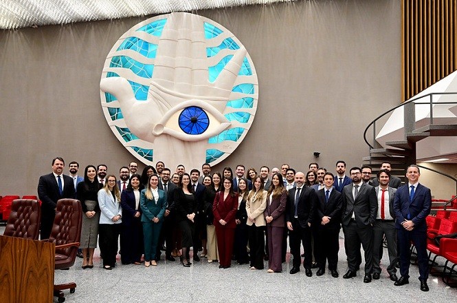 A imagem mostra um grande grupo de pessoas posando para uma foto em um ambiente interno, que parece ser uma sala de conferências ou um auditório. No primeiro plano, há cerca de 40 pessoas, todas vestidas de maneira formal, em trajes sociais. Elas estão em pé, agrupadas no centro da imagem e parecem estar sorrindo, o que sugere um momento de confraternização ou comemoração. Ao fundo, destaca-se uma grande escultura ou mural na parede que representa uma mão aberta com um olho no centro, adornada com detalhes que lembram águas em tons de azul e verde. Essa arte é a peça central da decoração e ocupa uma posição de destaque na sala. O ambiente tem um teto alto e há cadeiras vermelhas visíveis parcialmente nos cantos da imagem, o que reforça a sensação de estarmos em um espaço de eventos ou apresentações. Uma escada em espiral metálica está visível no lado direito, adicionando um elemento moderno ao cenário. A expressão das pessoas e a presença do traje formal sugerem um evento profissional ou institucional, possivelmente uma formatura, reunião ou posse. A atmosfera parece ser de celebração e solenidade. Quanto à época do dia ou estação, não há luz natural visível para uma definição clara, mas o ambiente interno é bem iluminado de forma artificial. A imagem é realista e capturada por uma câmera, apresentando detalhes nítidos dos participantes e do ambiente.