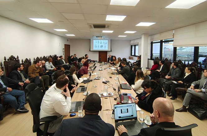 A imagem mostra uma reunião em uma sala de conferências. No primeiro plano, há várias pessoas sentadas ao redor de uma mesa longa de madeira, usando laptops e papéis. Elas parecem estar em uma discussão ou apresentação, com um projetor exibindo informações na tela ao fundo.