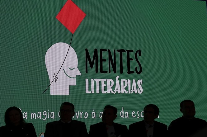 Imagem de um painel com a frase "MENTES LITERÁRIAS" em letras pretas sobre fundo verde.  Um desenho de uma cabeça humana em tons de cinza está presente no painel. Há também silhuetas de pessoas em frente ao painel, em trajes escuros.  Há outras frases em letras pretas sobre o fundo, indicando a frase "a magia do livro à escola".