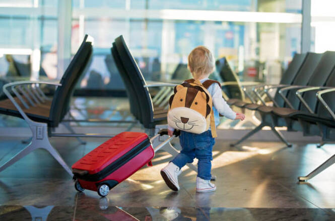 No primeiro plano da imagem, há uma criança pequena caminhando em um aeroporto. Ela está puxando uma mala vermelha de rodinhas e usa uma mochila com formato de animal, possivelmente um cachorro ou leão, o que adiciona um toque divertido ao cenário. Ao fundo, é possível ver cadeiras enfileiradas típicas de uma sala de espera de aeroporto, sugerindo que este é o local retratado. O ambiente parece ser iluminado pela luz do dia, possivelmente durante a manhã ou à tarde, devido à claridade. A criança está de costas, caminhando com confiança, o que transmite uma sensação de aventura e curiosidade. O contraste entre o pequeno tamanho da criança e a grande estrutura do aeroporto pode evocar emoções de ternura e fascínio. O ambiente tem uma atmosfera animada e de expectativa comum em aeroportos, sugerindo viagens e novas experiências. A imagem parece realista, capturando um momento cotidiano de maneira viva.