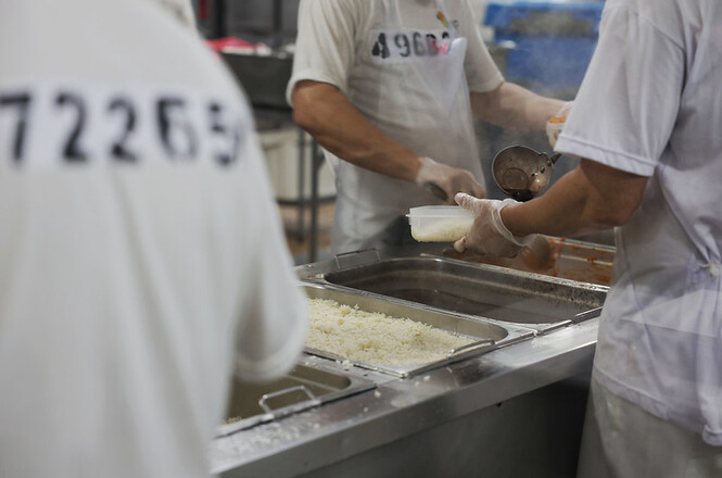 A imagem mostra uma cena em um estabelecimento provavelmente ligado a um refeitório ou cozinha industrial. No primeiro plano, há duas pessoas vestindo uniformes brancos, com números estampados nas costas das camisetas. Elas parecem estar servindo comida, utilizando colheres e vasilhames.