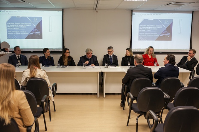 A imagem retrata uma reunião em uma sala de conferências. No primeiro plano, vemos várias pessoas sentadas ao redor de uma mesa de conferência branca. O grupo à frente da sala parece estar participando de uma discussão formal. Todos estão vestidos de maneira formal, usando ternos e roupas profissionais. Algumas pessoas estão fazendo anotações ou gesticulando enquanto falam. No fundo, há dois projetores mostrando o que parece ser uma apresentação de slides, o que sugere que o encontro é uma apresentação ou uma discussão de trabalho. A sala possui paredes brancas e um piso de madeira clara, criando um ambiente corporativo comum em escritórios ou universidades