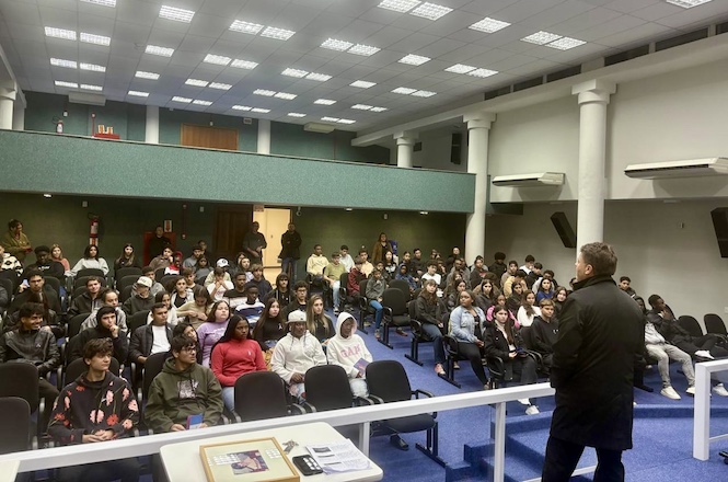 À direita, há um homem em pé, de costas para a câmera, aparentemente falando para a audiência. Ele veste um casaco preto. O auditório está cheio de pessoas sentadas em cadeiras voltadas para o orador. Há uma variedade de pessoas, algumas usando roupas casuais, outras com agasalhos. As expressões indicam atenção ao que está sendo dito. Há uma mesa na frente do orador com alguns papéis e objetos sobre ela. O auditório tem paredes verdes e um teto com iluminação artificial. As pessoas estão focadas no que o orador está dizendo, sugerindo um ambiente de palestra ou apresentação. A atmosfera parece ser de concentração e interesse, típica de uma situação de aprendizado ou apresentação. O ambiente é típico de uma sala de conferências ou auditório educacional. O uso de luz artificial sugere que a imagem pode ter sido tirada durante a noite ou em um espaço fechado sem luz natural direta. A imagem é realista e mostra uma cena cotidiana que parece não ser modificada ou gerada por computador.