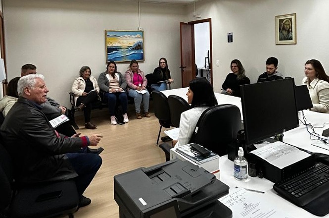 A imagem retrata um escritório com várias pessoas sentadas em um círculo, possivelmente participando de uma reunião ou discussão. No primeiro plano, há uma mesa com computadores, um telefone, papéis e um frasco de álcool em gel. No fundo, sentadas em cadeiras ao longo das paredes, há pessoas que parecem ouvir ou interagir umas com as outras.