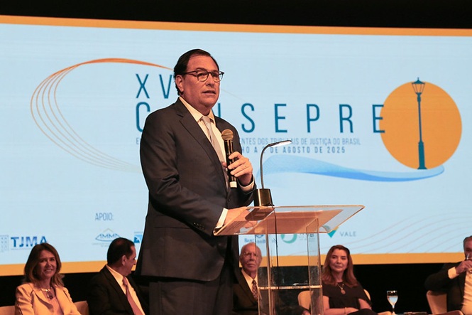 Foto horizontal: Francisco Oliveira Neto discursa ao microfone em congresso jurídico com palco iluminado em azul.