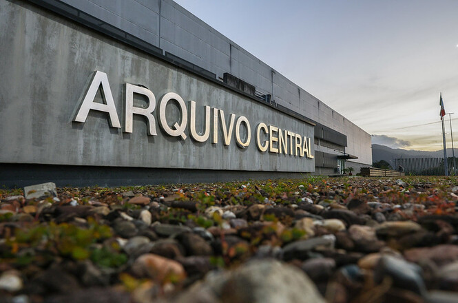 A imagem mostra um edifício moderno identificado como "ARQUIVO CENTRAL". No primeiro plano, pode-se ver o solo coberto de pequenas pedras e vegetação rasteira. No fundo, aparece uma estrutura arquitetônica com uma fachada de concreto, complementada por grandes letras em relevo que formam o nome da instituição.O ambiente ao redor parece ser calmo e organizado, típico de uma área urbana destinada ao uso institucional ou administrativo. No plano de fundo, é possível avistar algumas bandeiras, sugerindo a presença de um edifício oficial ou governamental.
