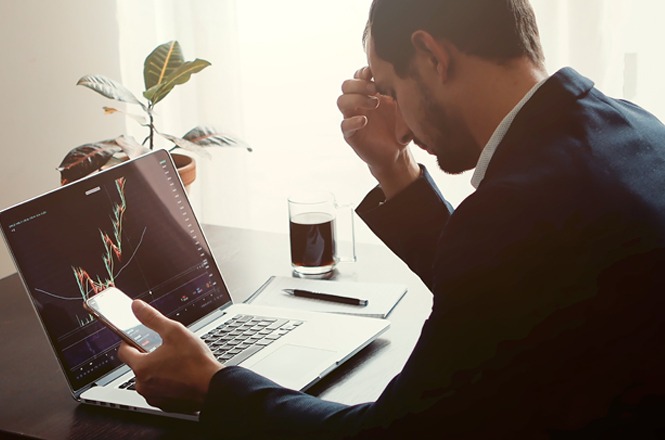 No primeiro plano, está um homem de terno, segurando um celular com uma mão e apoiando a cabeça com a outra, em frente a um laptop.