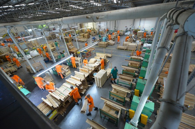A imagem mostra o interior de uma fábrica de carpintaria, com pessoas trabalhando entre bancadas cheias de tábuas e materiais de madeira.