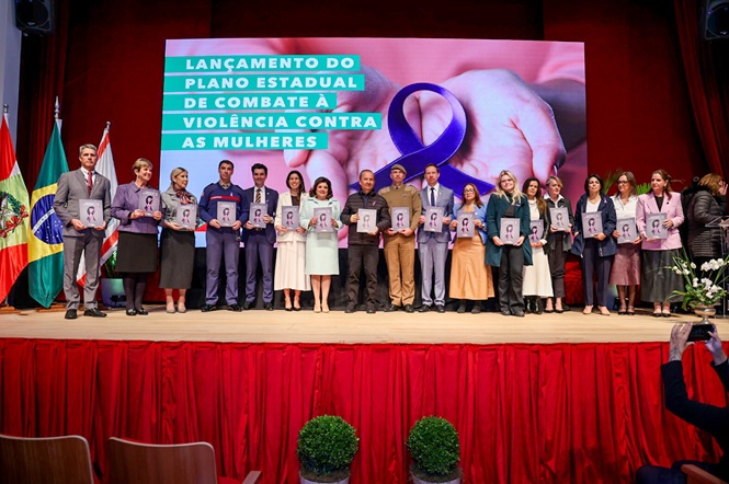 A imagem mostra um grupo participando do “Lançamento do Plano Estadual de Combate à Violência Contra as Mulheres”, com laço roxo símbolo da campanha.