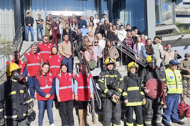 Um grupo posa em uma escadaria diante de um edifício, com destaque para bombeiros de uniforme e capacete amarelo e pessoas de colete vermelho com emblema.