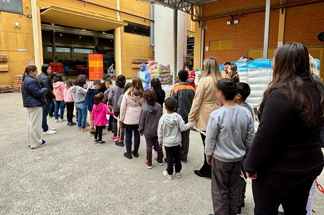 Uma grande quantidade de crianças, em fila, acompanhadas por adultos. As crianças estão de pé em fila indiana, e parecem estar em um ambiente externo, possivelmente um depósito ou galpão.